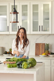 A woman smiling in a kitchen, surrounded by fresh vegetables, representing a healthy lifestyle aligned with WeNatal's prenatal supplement range.
